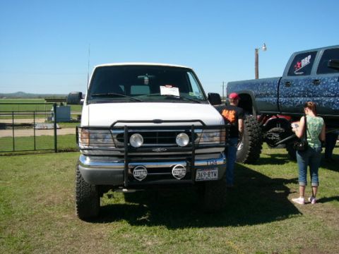 1978 Ford Bronco - Front view of a white lifted 4x4 Ford Econoline van parked on grass at an outdoor show, wearing a black brush guard with round driving ligh…