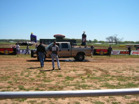 1978 Ford Bronco - A pickup truck is parked on the dirt track at a drag strip event with spectators and inflatable advertising in the background.