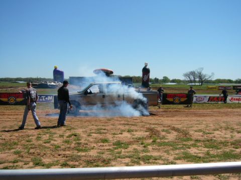 1978 Ford Bronco - A pickup truck performs a burnout on a dirt track, with spectators and large inflatable advertisements in the background.