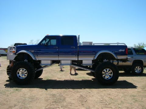 1978 Ford Bronco - Side profile of a heavily lifted blue and tan two-tone Ford crew cab pickup on a dirt show field, sitting on tall mud tires with chrome eig…