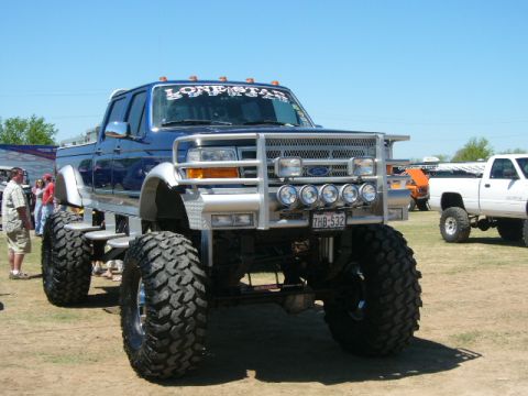 1978 Ford Bronco - Front three-quarter view of a heavily lifted blue and silver Ford crew cab pickup on a dirt show field, running huge mud-terrain tires and…