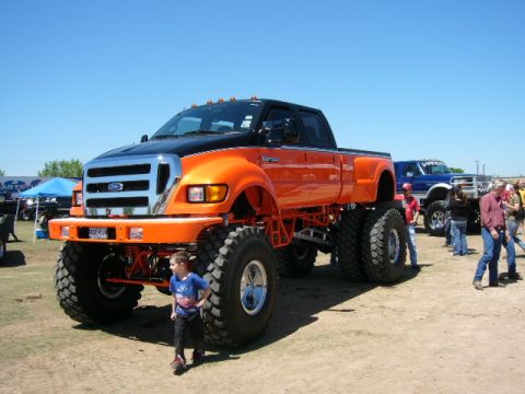 1978 Ford Bronco - Orange and black Ford F-650 crew cab pickup on a massive lift, photographed from the 3/4 front on a dirt show field. It rides on huge milit…