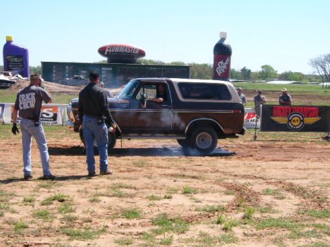 1978 Ford Bronco - Brown and tan two-tone full-size Ford Bronco with a white removable hardtop, shown in side profile doing a smoky burnout on a dirt track. T…