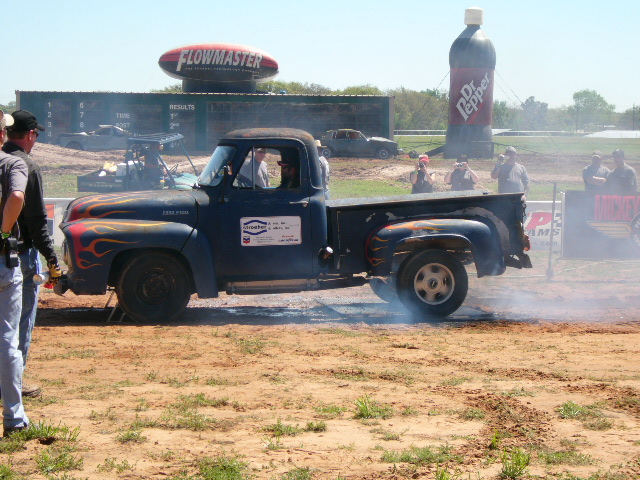 Old blue pickup truck with flames doing a burnout, smoke coming from tires. Flowmaster and Dr. Pepper signs in background