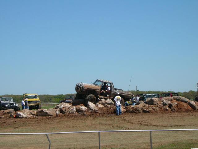 Mud-covered 1977 Ford F-150 pickup truck on rocks, front 3/4 view, with other vehicles in background