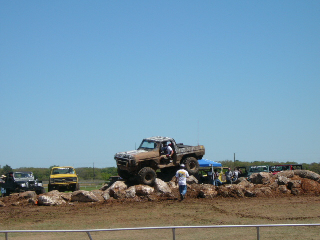 Brown off-road truck climbing over rocks. Another yellow truck and black jeep are parked nearby
