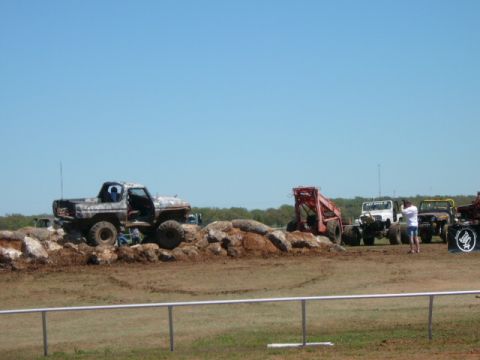 1978 Ford Bronco - Several off-road vehicles, including a skid steer loader, are positioned on a dirt track with rocky terrain under a clear blue sky.