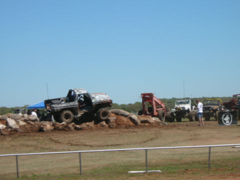 1978 Ford Bronco - Several off-road vehicles are gathered on a dirt track with rocks and trees in the background under a clear blue sky.