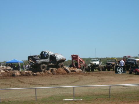 1978 Ford Bronco - Several off-road vehicles are gathered on a dirt course under a clear blue sky, with spectators visible near a blue tent.