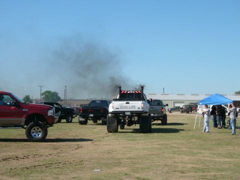 1978 Ford Bronco - A white pickup truck emits a large cloud of black smoke from its dual exhaust stacks, with other vehicles and people in the background.