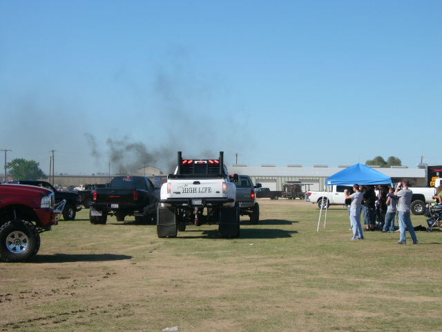Several modified pickup trucks are parked on a dirt field, one is emitting a large cloud of black smoke. People are gathered under a blue tent