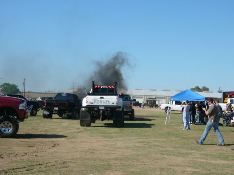 1978 Ford Bronco - Several trucks are gathered on a grassy field under a clear blue sky, with one truck producing a large plume of black smoke.