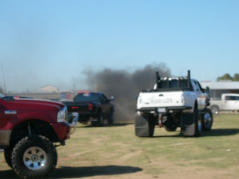 1978 Ford Bronco - A white pickup truck with large tires and exhaust stacks emits a large cloud of black smoke, with other trucks visible in the background.