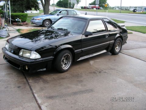 1988 Ford Mustang - A black coupe is parked on a concrete driveway with a light blue sedan in the background.