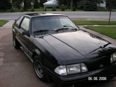 1988 Ford Mustang - Black coupe parked on a driveway with a street and trees in the background. The car is viewed from the front 3/4 angle.