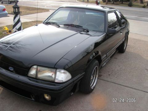 1988 Ford Mustang - A black coupe is parked on a concrete driveway with a street and trees in the background.