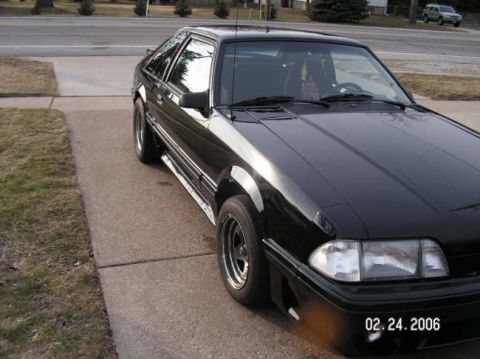 1988 Ford Mustang - A black coupe is parked on a concrete driveway with grass and trees in the background.