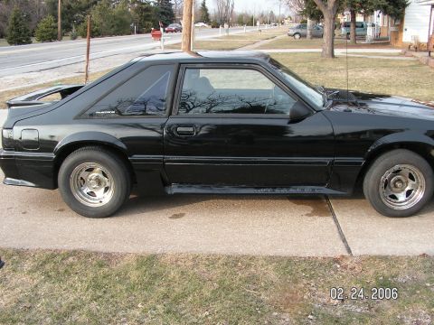 1988 Ford Mustang - A black coupe car is parked on a concrete driveway with grass and trees in the background.