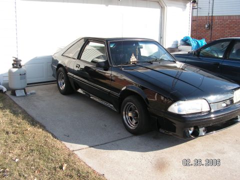 1988 Ford Mustang - A black coupe is parked in front of a garage, with another vehicle visible to its right.