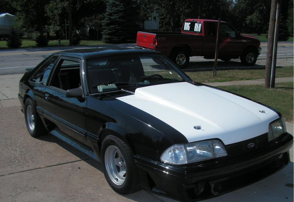 Black and white Ford Mustang with a white hood and hood pins, parked on a driveway. A red pickup truck is visible in the background