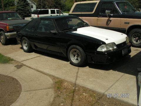 1988 Ford Mustang - A black 1988 Ford Mustang with a white hood is parked in a driveway with other vehicles.