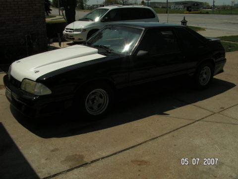 1988 Ford Mustang - A black coupe with a contrasting white hood sits parked on a concrete driveway, with a white minivan in the background.