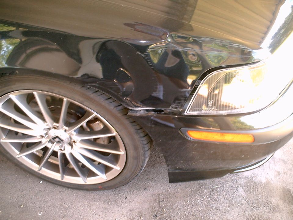 Front-left corner of black sedan with bumper damage and aftermarket wheel