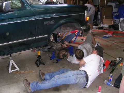 1994 Ford Bronco - Several people are gathered around a lifted Ford Bronco, appearing to work on its front axle and suspension components.