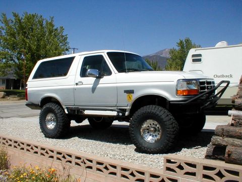 1996 Ford Bronco - Three-quarter front view of a lifted white Ford Bronco on tall mud-terrain tires with polished aluminum wheels, fitted with a black tubular…