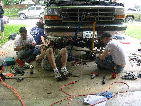 1994 Ford Bronco - Mechanics work on the front end of a 1994 Ford Bronco, with another SUV and pickup truck visible in the background.