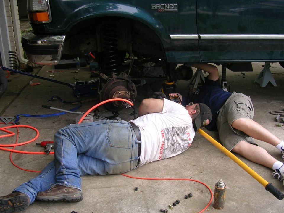 Two men working on the undercarriage of a green 1994 Ford Bronco, with tools and parts scattered around