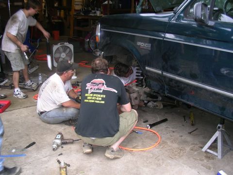 1994 Ford Bronco - Several people are gathered around a dark green Ford Bronco, appearing to work on its suspension in a garage setting.