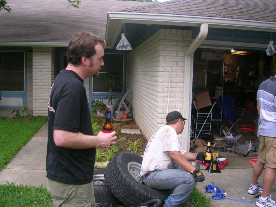 Two men with beer bottles stand near stacked tires and a garage