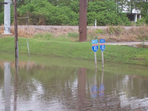 1992 Ford Bronco - A flooded road with two highway signs indicating I-10 West and East, surrounded by greenery and utility poles.