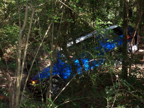1992 Ford Bronco - A blue SUV has gone off-road and is resting on its side amongst dense foliage.