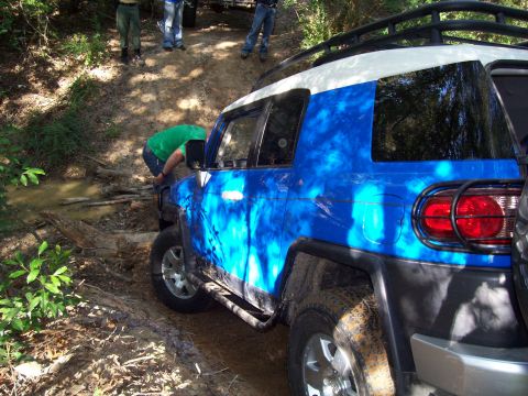 1992 Ford Bronco - A group of people attempt to free a blue and white SUV stuck in a muddy off-road trail.