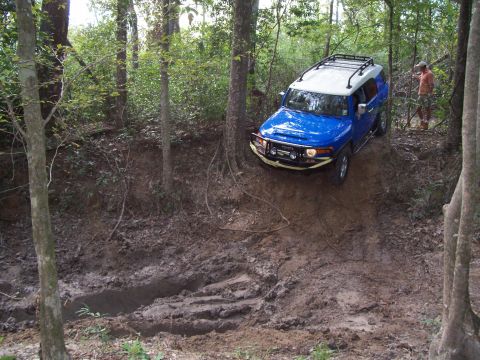 1992 Ford Bronco - A blue Toyota FJ Cruiser navigates a challenging muddy path through a wooded area.