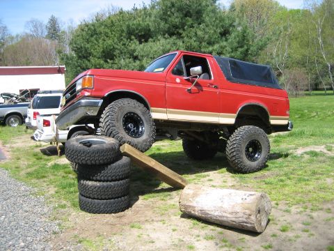 1990 Ford Bronco - Lifted red-and-tan full-size Ford Bronco with a black soft top flexing its suspension as it climbs a makeshift ramp of stacked mud tires, a…