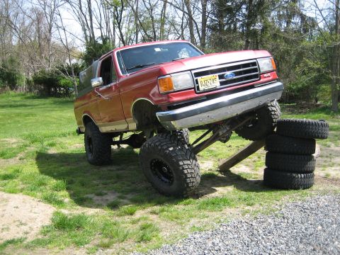 1990 Ford Bronco - Red and tan lifted Ford Bronco parked on a grass yard with its passenger front wheel ramped up on a wooden plank next to a stack of four sp…