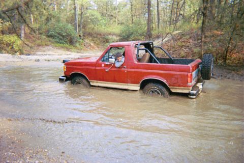1990 Ford Bronco - A red 1990 Ford Bronco with a roll cage navigates through a muddy water crossing.