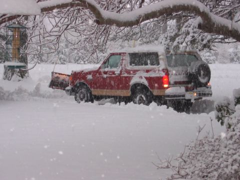 1990 Ford Bronco - Red-and-tan full-size Ford Bronco with a mounted snow plow blade pushing snow through a driveway during heavy snowfall, seen from the passe…