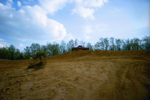 1990 Ford Bronco - A red pickup truck is parked on top of a dirt mound under a cloudy sky.