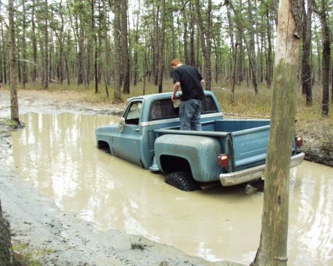 1989 Ford Bronco - A light blue Chevrolet C/K pickup truck is stuck in a muddy forest road, with a person standing in the truck bed.