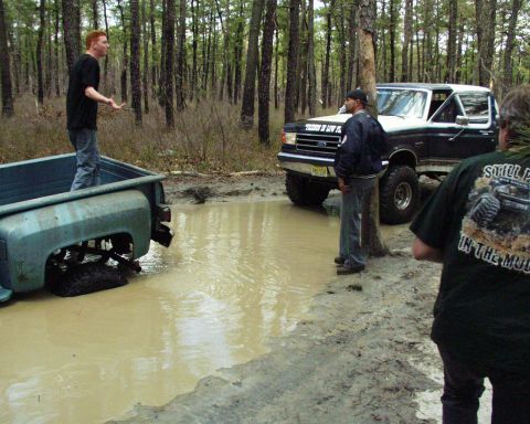 1989 Ford Bronco - Two trucks are stuck in a muddy puddle in a wooded area.