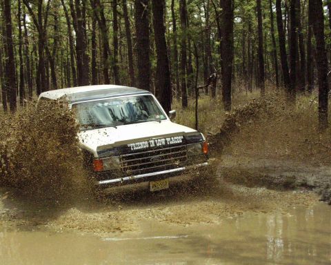 1989 Ford Bronco - A white 1989 Ford Bronco navigates a muddy trail, kicking up a large spray of water and dirt.