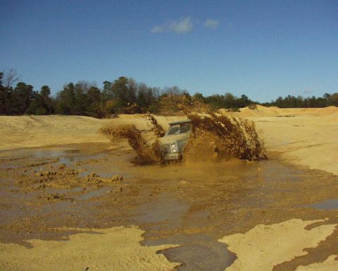 1989 Ford Bronco - A light-colored SUV navigates through a muddy terrain, kicking up a large spray of dirt and water.