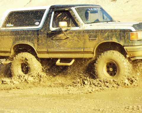 1989 Ford Bronco - A muddy Ford Bronco churns through a dirt track, kicking up a large spray of mud.