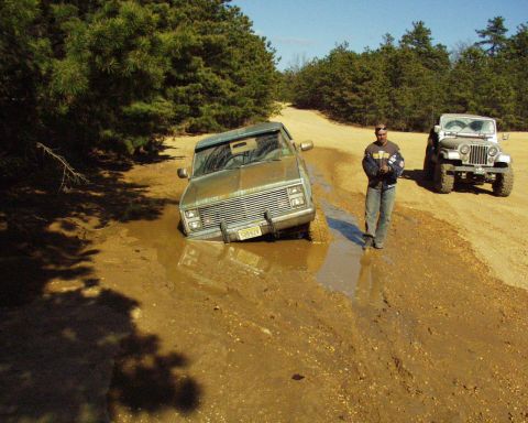 1989 Ford Bronco - A man stands by a muddy road as a van is stuck in a large mud puddle, with a Jeep in the background.
