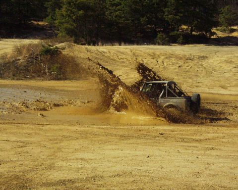 1989 Ford Bronco - An off-road vehicle navigates a muddy terrain, kicking up a large spray.