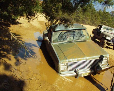 1989 Ford Bronco - A mud-covered pickup truck is stuck in a muddy puddle, with a Jeep visible in the background.